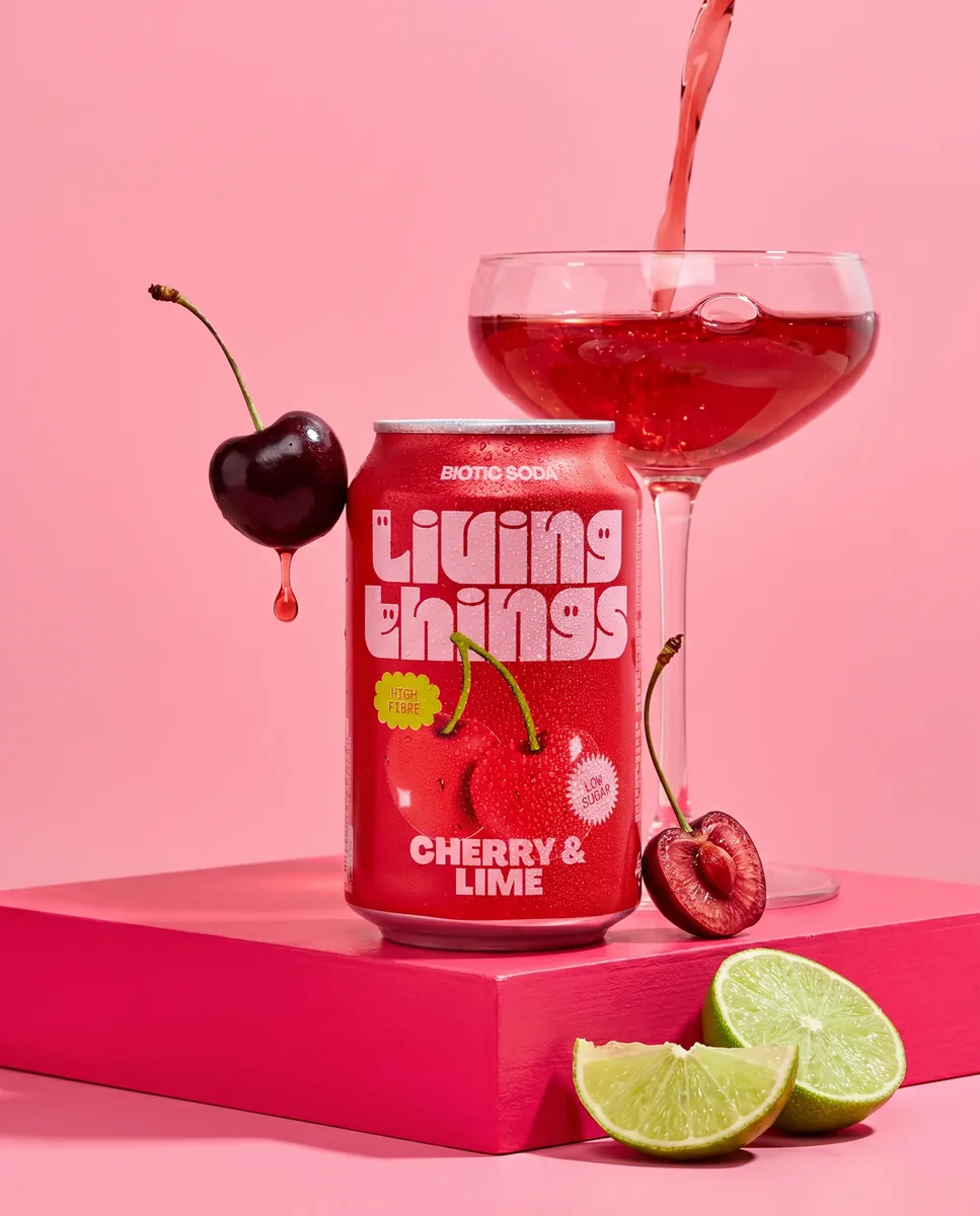 Hero image of a cherry and lime soda can on a pink pedestal beside a coupe glass mid-pour with a floating cherry