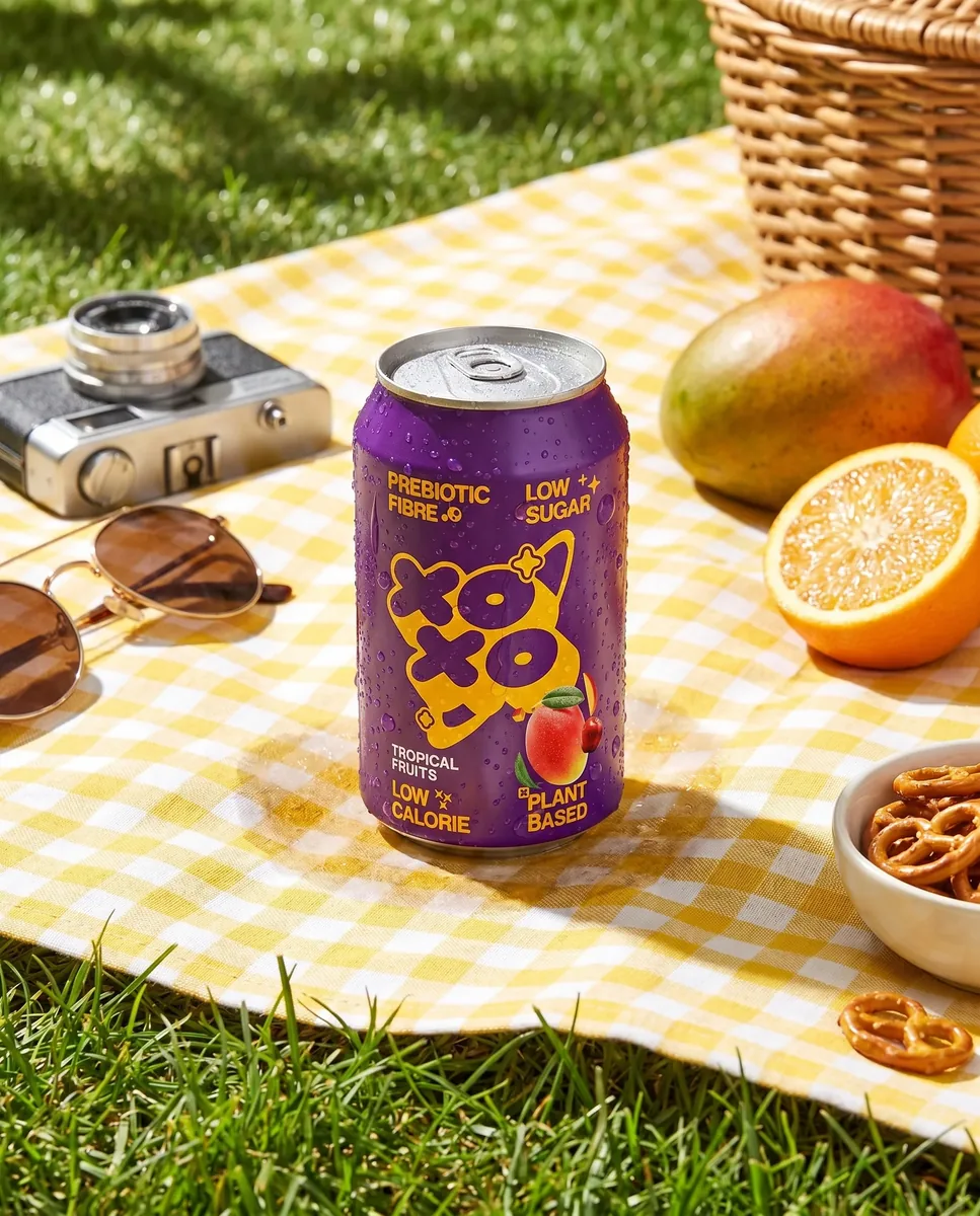 A soft drink can on a yellow gingham picnic blanket beside a vintage camera, sunglasses and fresh fruit
