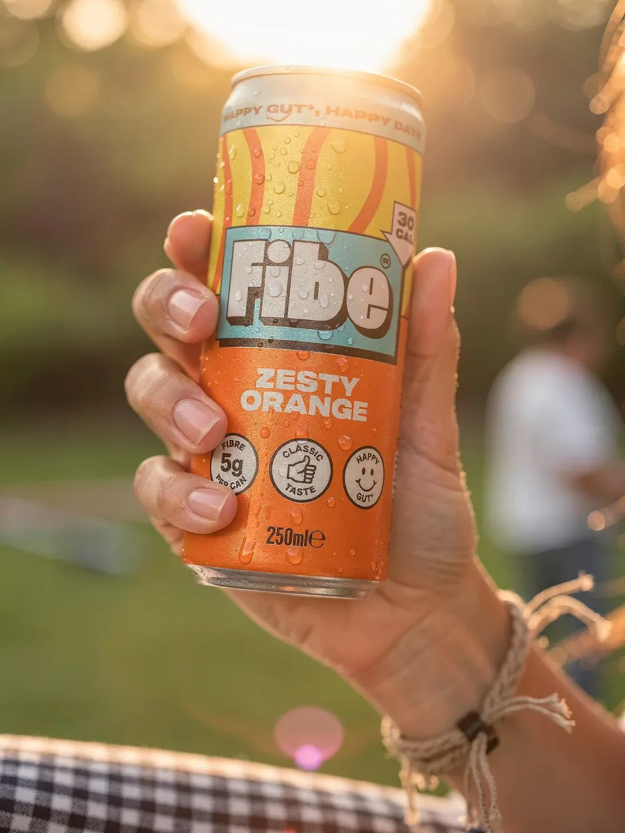 A hand holding a tall orange soft drink can outdoors at sunset with warm backlight