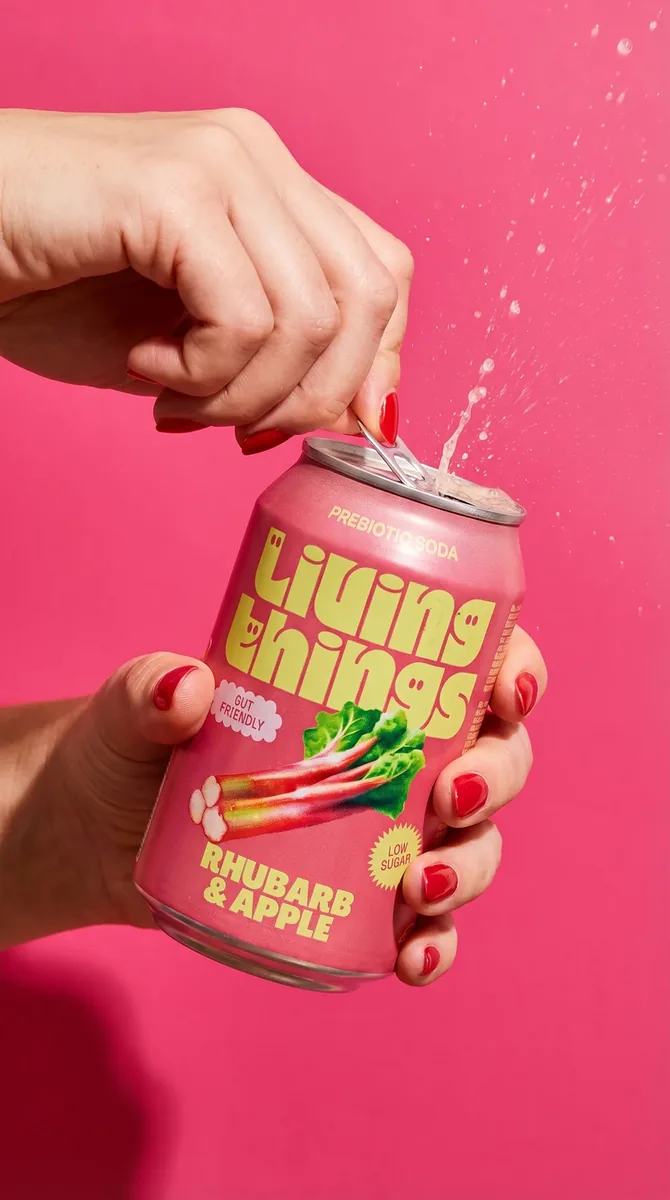 A hand cracking open a soft drink can with liquid spraying from the ring-pull against a hot pink backdrop